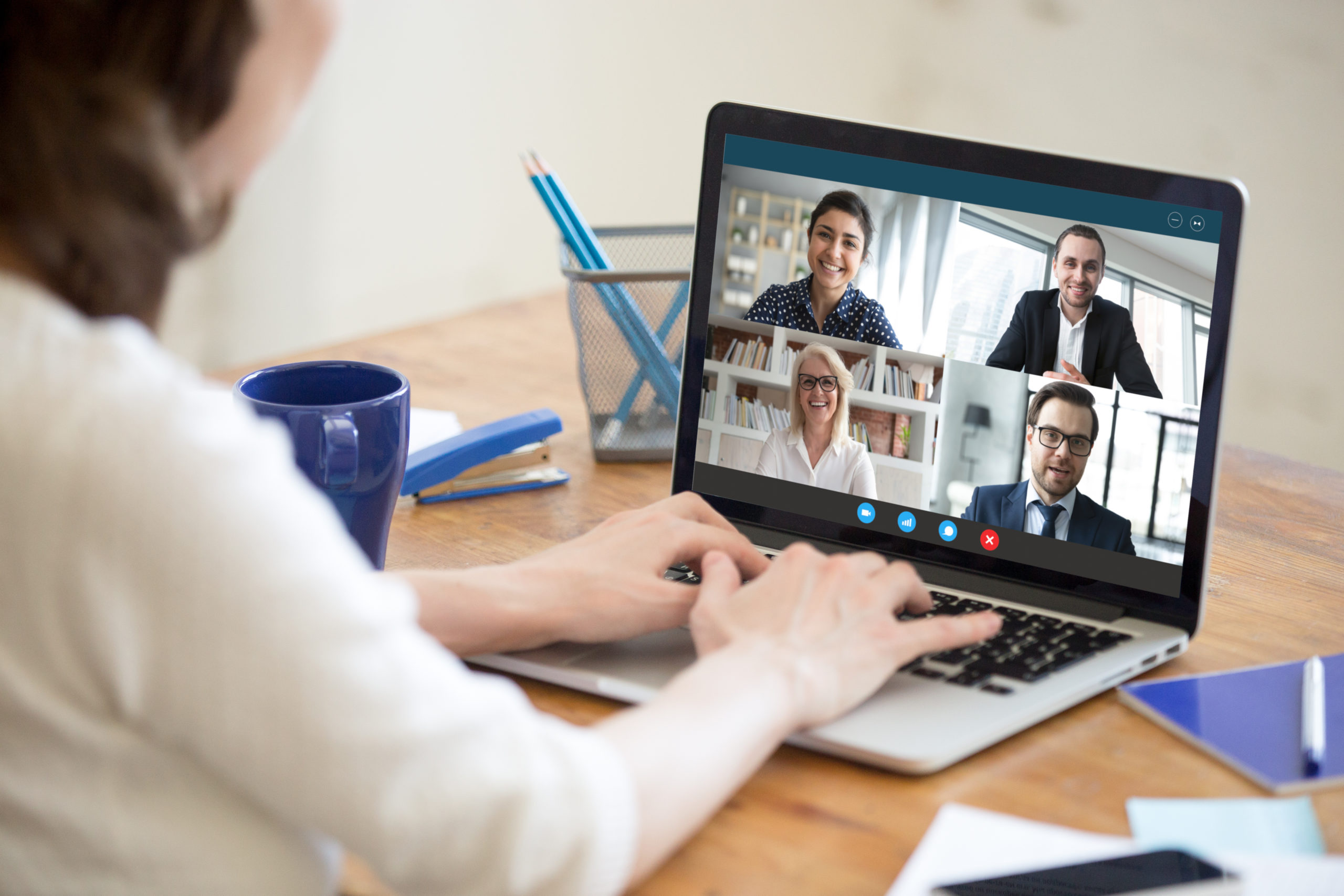 Diverse people take part in group video call pc screen cam app view over woman shoulder, seated at desk. Solve business issues distantly during coronavirus pandemic outbreak, videoconferencing concept